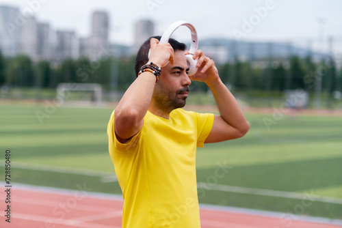 Man in yellow putting on wireless headphones to exercise with inspiring music at outdoor city sports stadium with empty football field sportsman enjoying workout photo