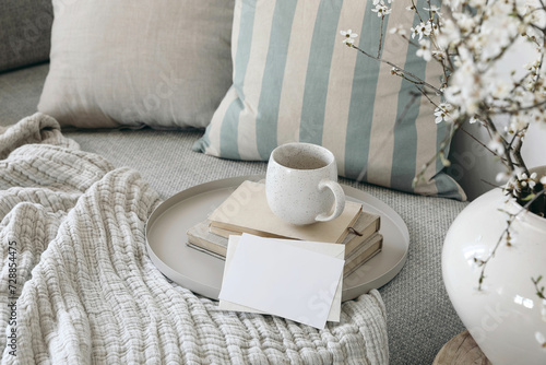 Spring breakfast scene. Blank greeting card mockup. Cup of coffee, tea on books. Round beige tray. Blossoming cherry plum tree branches in ceramic vase. Cozy linen sofa, cushions. Easter still life.