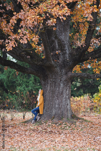 A beautiful blonde woman is standing by the wide old oak tree in autumn.  © Konstantins Pobilojs