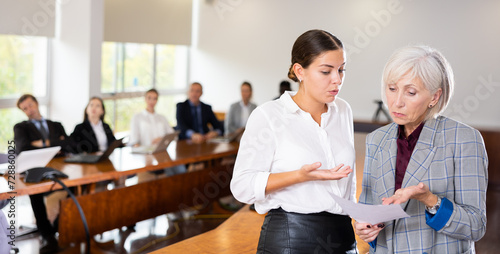 Portrait of two age-diverse white female colleagues standing in front of other coworkers in conference room and discussing business report during team meeting at office