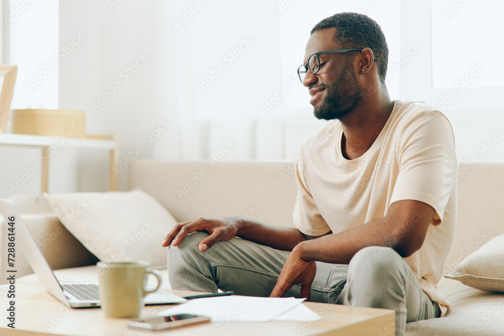Smiling African American Freelancer Working on Laptop in Modern Home Office A young African American man is sitting on a comfortable sofa in a stylish living room He is focused and smiling, typing on