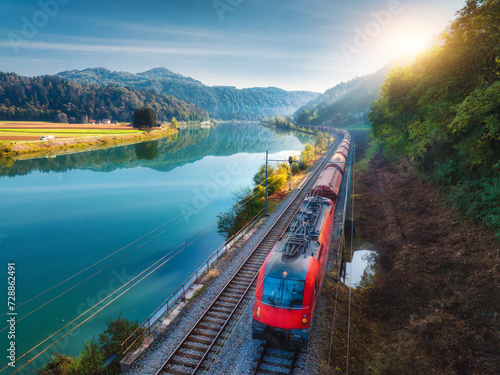 Aerial view of red modern high speed train moving near river in alpine mountains at sunrise in spring. Top view of train, railroad, lake, reflection, green trees in summer. Railway station in Slovenia