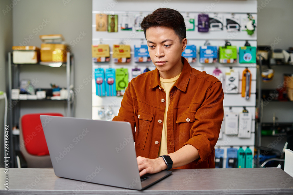 serious asian man using laptop on counter in private electronics store, small business concept serious asian man using laptop on counter in private electronics store, small business concept