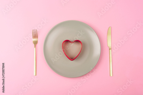 Gray plate with red cookie cutter in a shape of heart, fork and knife on pink background. Valentines day concept. Top view, flat lay