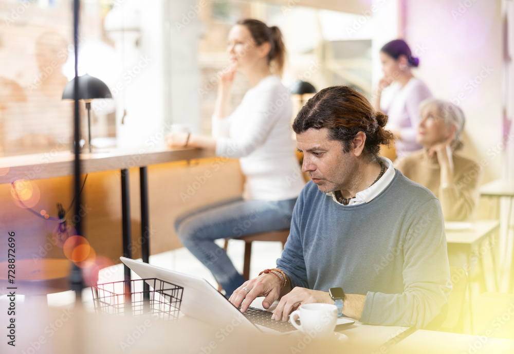 Fototapeta premium Concentrated male designer preparing new project on laptop, sitting in cozy cafe with cup of coffee