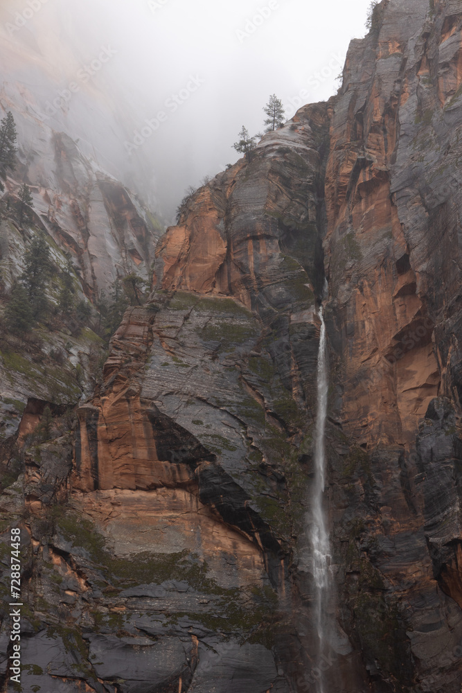 An ephemeral waterfall pours down the wet red and black sandstone cliffs of Zion Nat. Park in Utah, USA while low clouds fill the canyon above.
