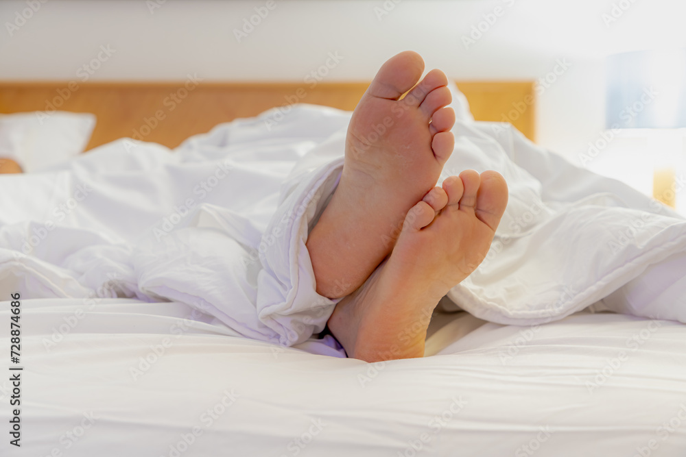 Selective focus of a men feet lied down on bed, Relaxing and watching TV on white bed sheet, A man showing his bare foot on blanket with lamp light at night in bedroom. Selective focus of a men feet lied down on bed, Relaxing and watching TV on white bed sheet, A man showing his bare foot on blanket with lamp light at night in bedroom.