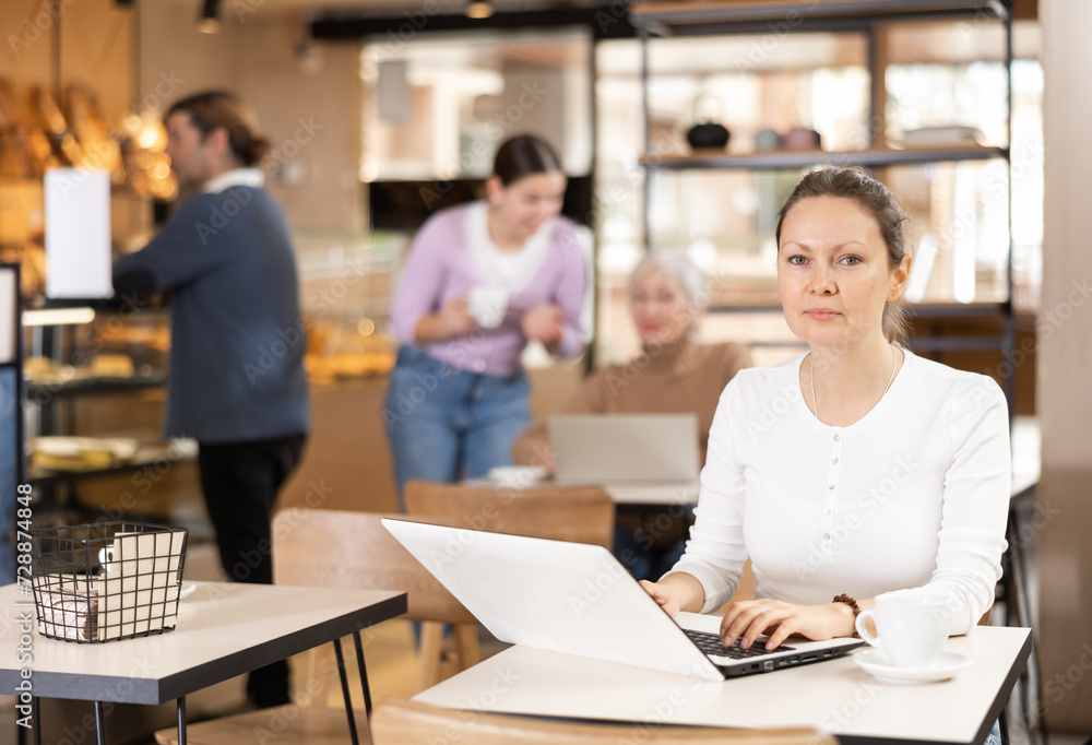 Fototapeta premium Focused woman drinking coffee while working on laptop in cozy cafe