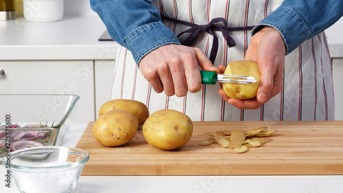 Closeup of man in an apron peeling potatoes over a kitchen counter. (ID: 728875600)