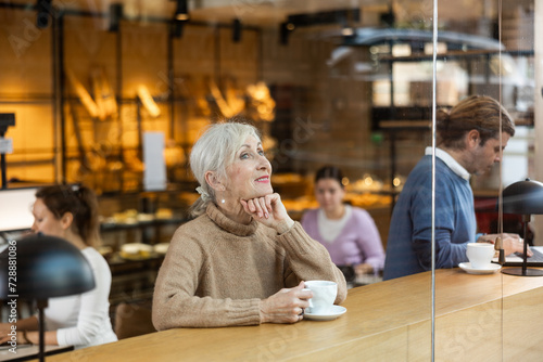 Smiling mature female in casual wear sitting at table and enjoying coffee or tea in cozy cafeteria