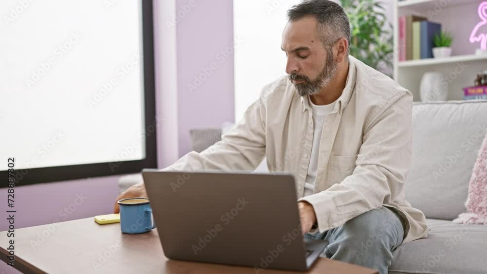 A mature man with a beard and grey hair sits on a sofa at home, working on a laptop and talking on the phone. A mature man with a beard and grey hair sits on a sofa at home, working on a laptop and talking on the phone.