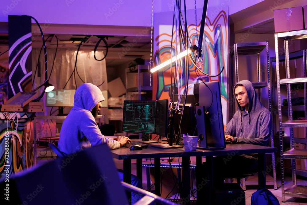 Naklejka premium Internet criminals working together to break into database in dark room. African american and asian young men hacking computer system and installing virus malicious software