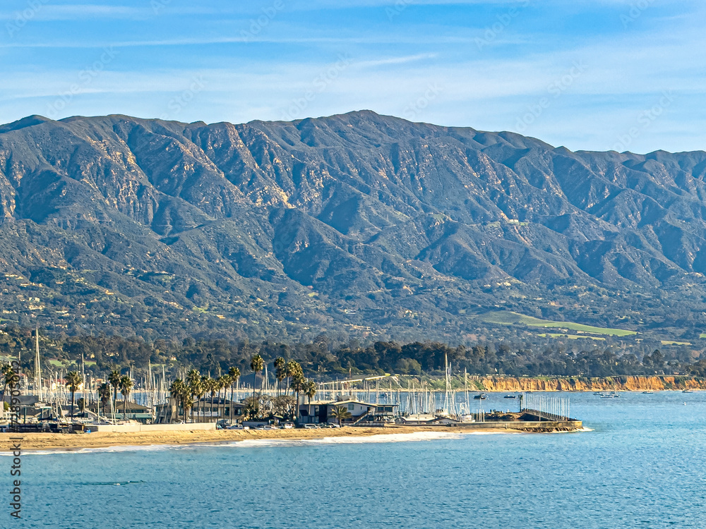 Santa Barbara, CA, USA - January 28, 2024: Harbor south side with harbor Walk pier and Castillo Point on corner. Collection of white boat masts. Mountain range in back under blue cloudscape