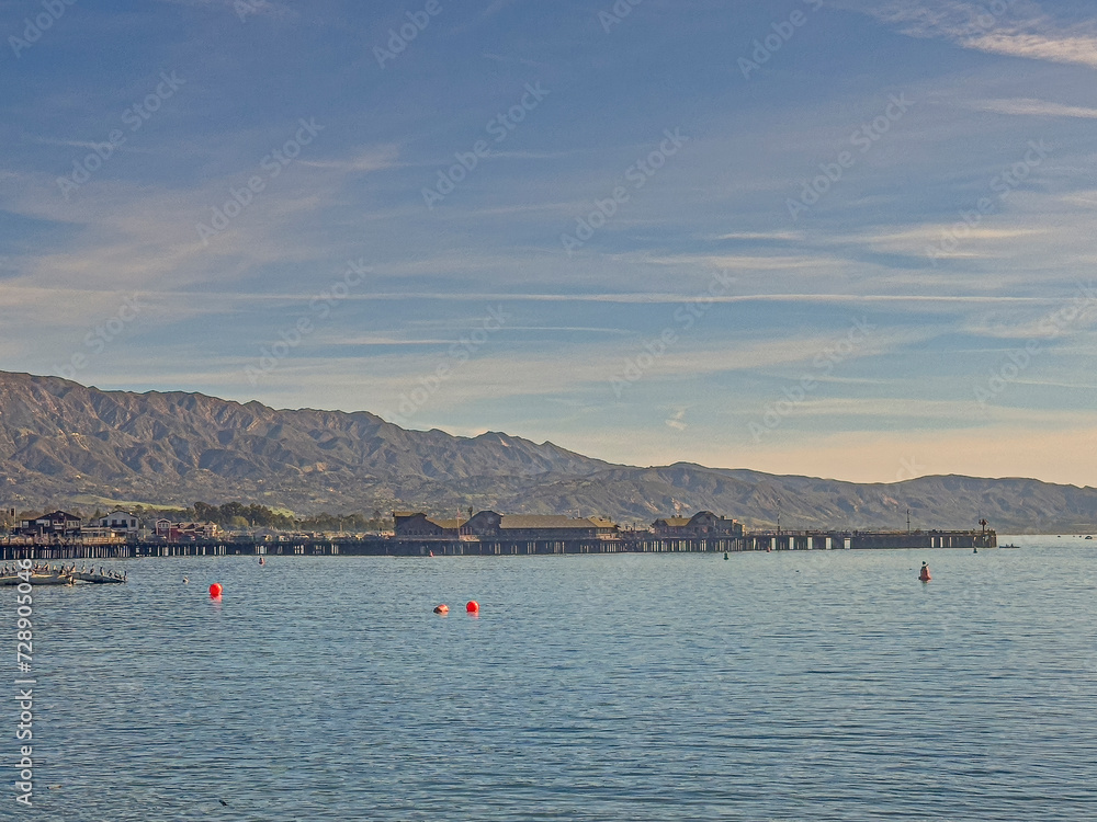 Santa Barbara, CA, USA - January 28, 2024: Entire length of Stearns Wharf pier with buildings on top. Wide landscape with ocean up front, mountain range, all under blue morning cloudscape
