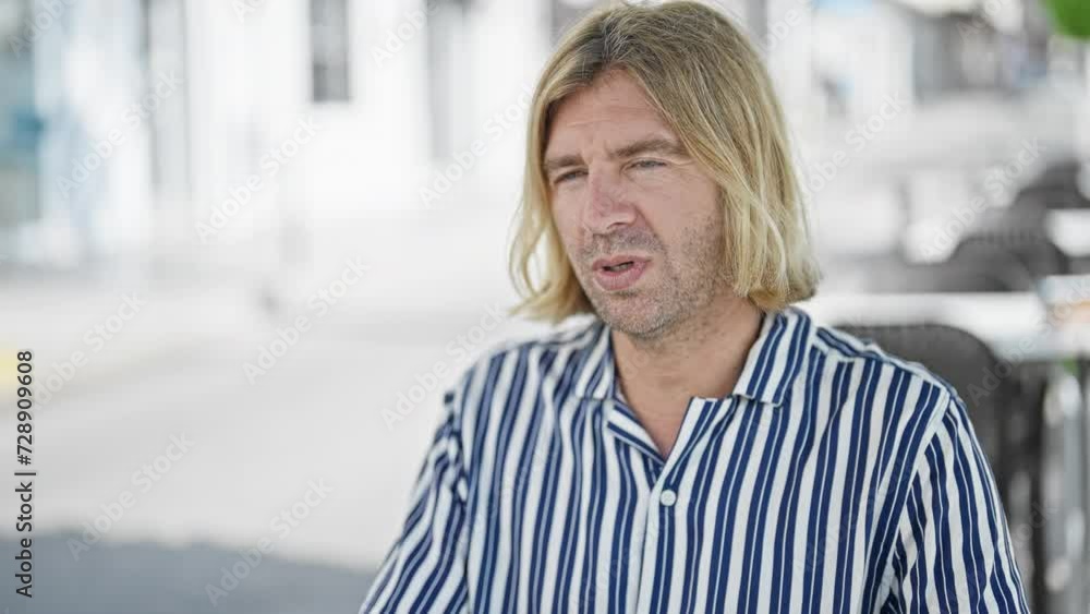 A middle-aged man with blond hair, casual striped attire, sitting thoughtfully at a cafe terrace in an urban setting. A middle-aged man with blond hair, casual striped attire, sitting thoughtfully at a cafe terrace in an urban setting.