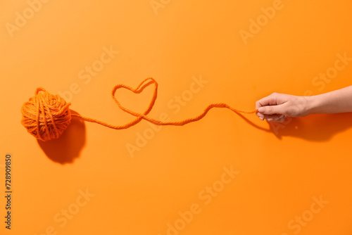 Female hand holding thread of yarn ball with heart on orange background