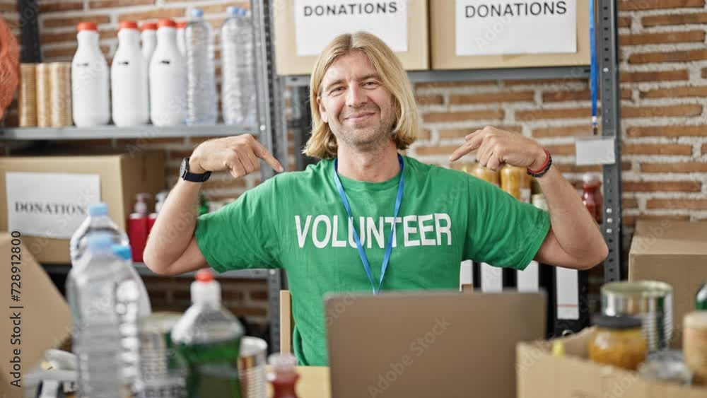 Smiling blond man wearing a volunteer shirt working on a laptop in a donation center warehouse with shelves of supplies in the background. Smiling blond man wearing a volunteer shirt working on a laptop in a donation center warehouse with shelves of supplies in the background.