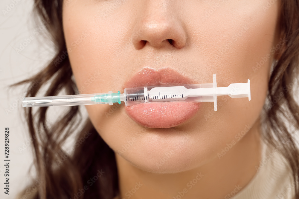 Young woman holding injection syringe in mouth on grey background