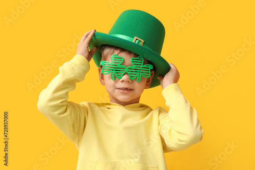 Cute little boy in leprechaun hat and decorative glasses in shape of clover on yellow background. St. Patrick's Day celebration