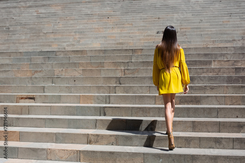 Mujer delgada de pelo largo y vestido amarillo subiendo escaleras bajo el sol photo