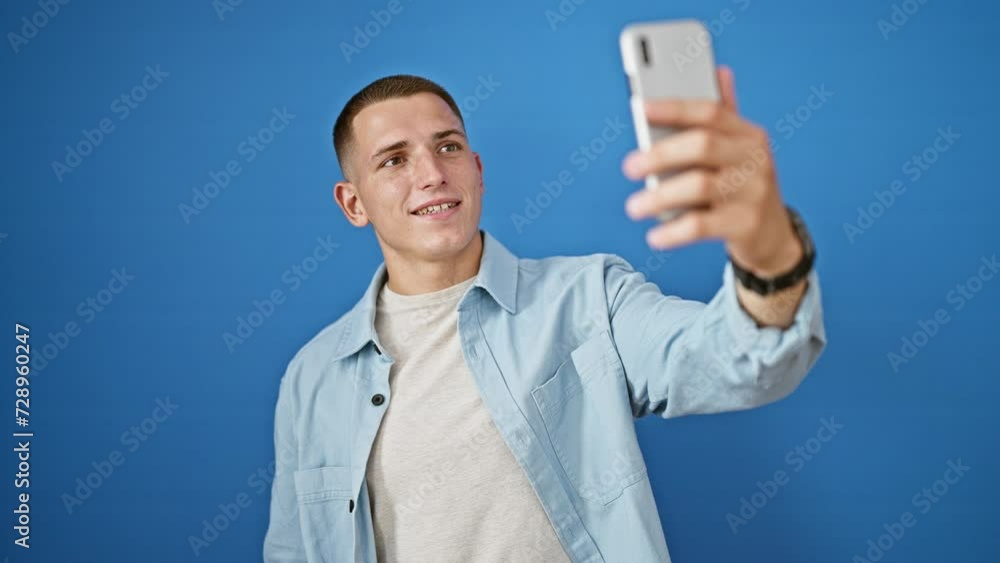 A smiling young hispanic man takes a selfie with his smartphone against an isolated blue wall outdoors. A smiling young hispanic man takes a selfie with his smartphone against an isolated blue wall outdoors.