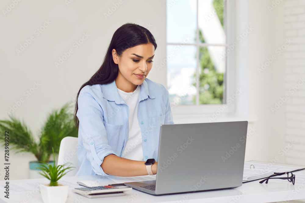 Confident young business woman accountant wearing casual clothes sitting at the desk on her workplace, working on a laptop in modern office and looking at the computer monitor screen. Confident young business woman accountant wearing casual clothes sitting at the desk on her workplace, working on a laptop in modern office and looking at the computer monitor screen.