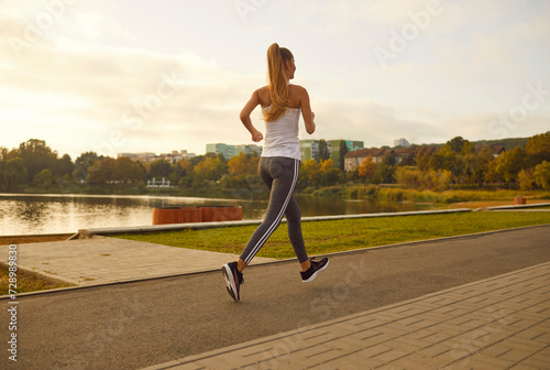 Back view of female young runner jogging alone in city park. Healthy sporty fitness woman running outdoors training for wellness, health and cardio. Workout in nature and healthy lifestyle concept.