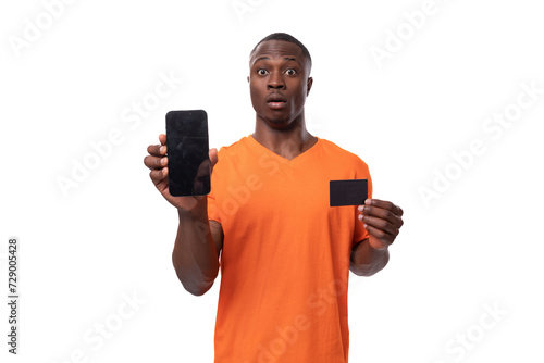 young surprised african man dressed in orange t-shirt holding phone and credit card. advertising concept with mockup