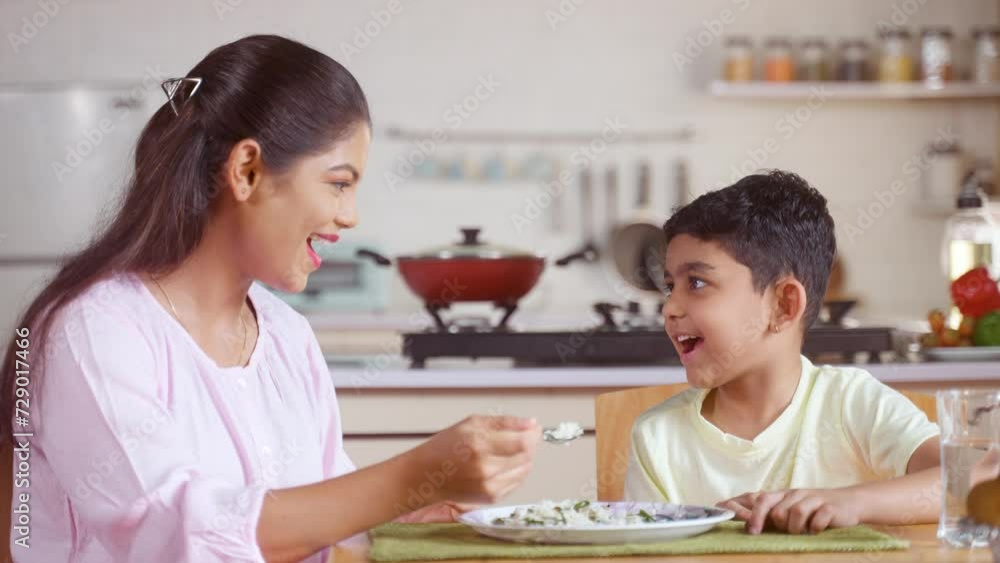 Happy Indian mother feeding to child at home on dining table - concept of parental caring, family bonding and motherhood. Happy Indian mother feeding to child at home on dining table - concept of parental caring, family bonding and motherhood.