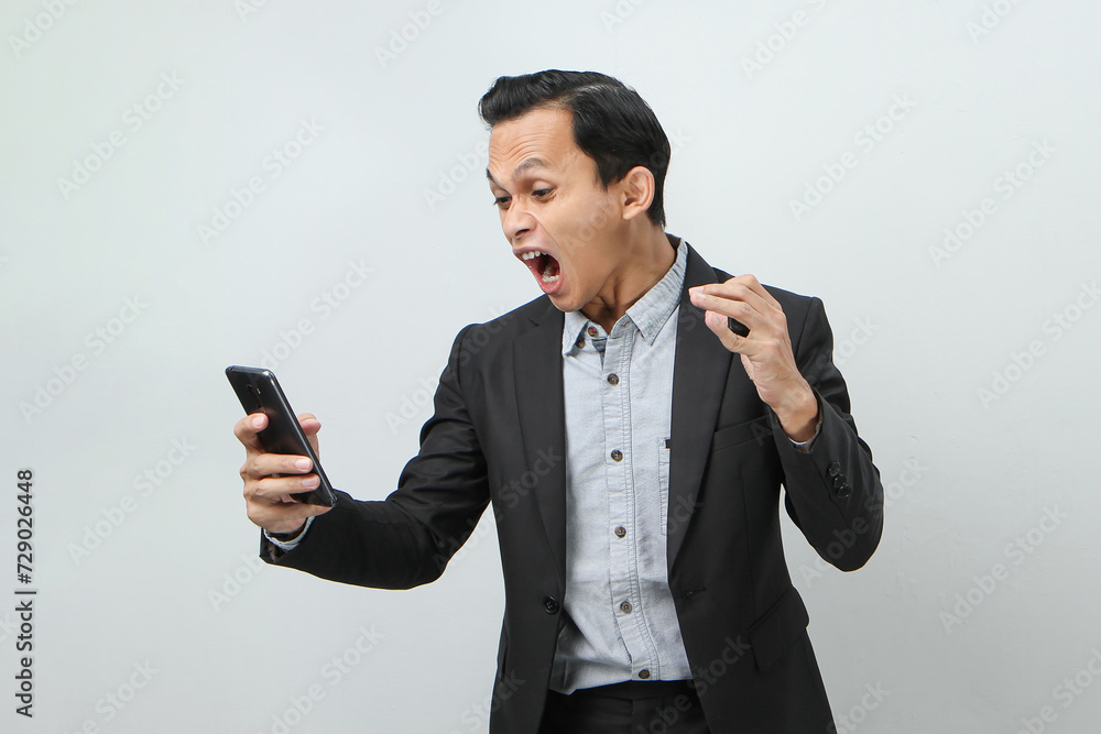 angry mad asian indonesian business man in suit holding smart phone on isolated background angry mad asian indonesian business man in suit holding smart phone on isolated background
