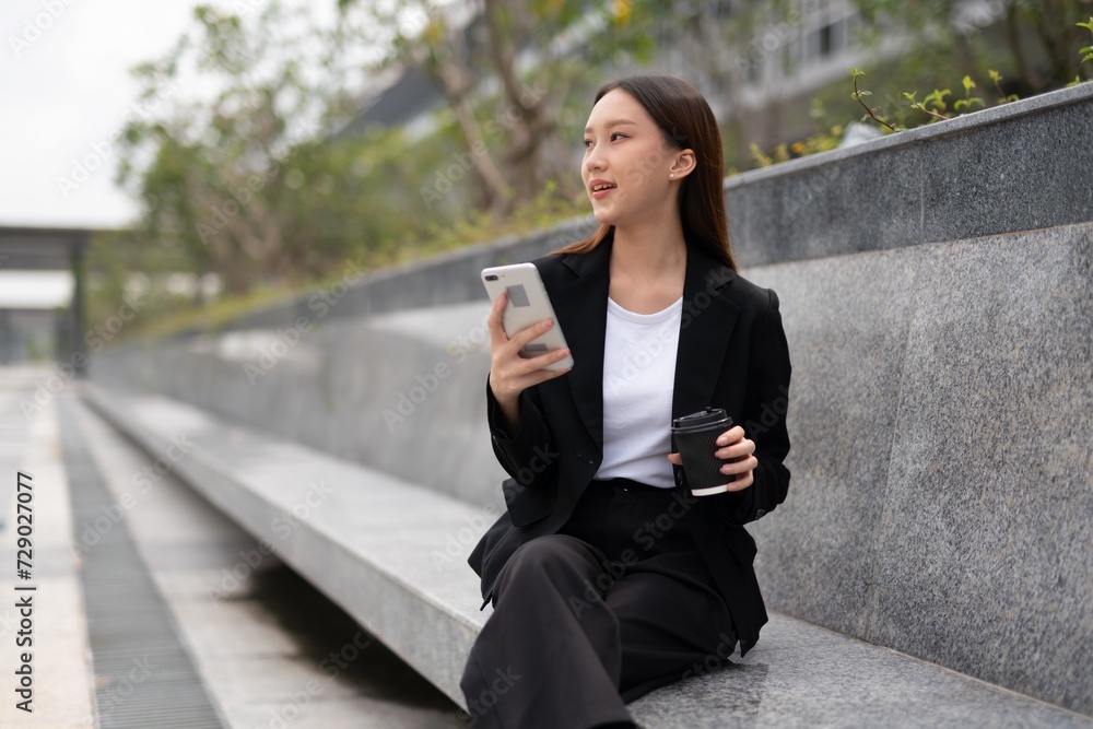 Young asian woman sitting and using phone outdoor, holding a cup of coffee. Business on the move. Taking a break. Young asian woman sitting and using phone outdoor, holding a cup of coffee. Business on the move. Taking a break.