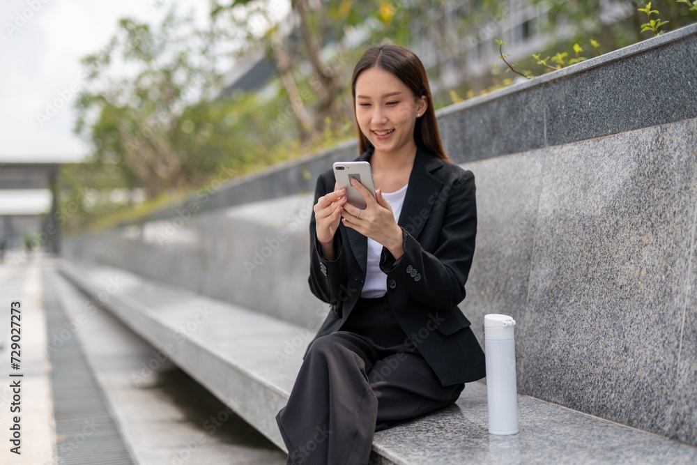 Asian businesswoman sitting and using phone outdoor, with a tumbler beside her Asian businesswoman sitting and using phone outdoor, with a tumbler beside her