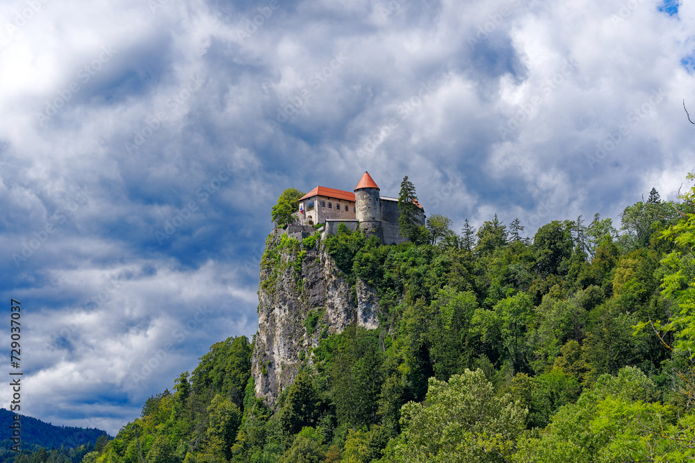 Castle on top of rock with waving Slovenian flag at lakeshore of Slovenian Lake Bled on a blue cloudy summer day. Photo taken August 8th, 2023, Bled, Slovenia.