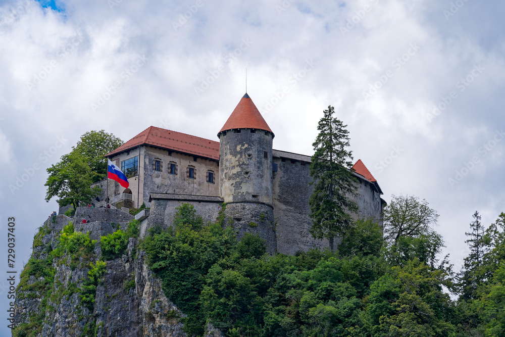 Scenic view of castle on top of rock with Slovenian flag waving at lakeshore of Slovenian Lake Bled on a cloudy summer day. Photo taken August 8th, 2023, Bled, Slovenia.