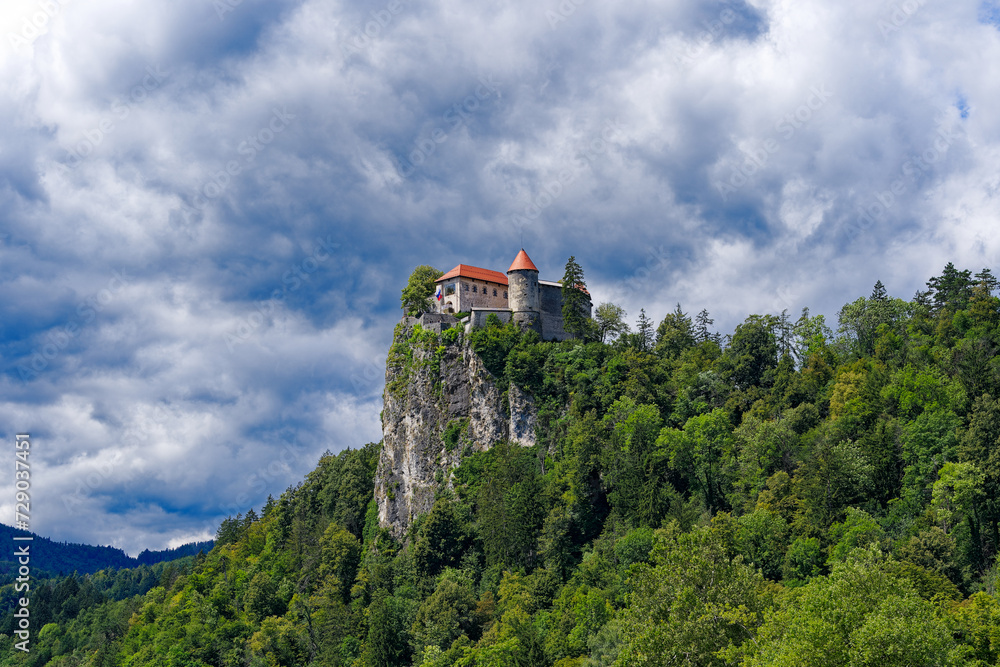 Scenic view of castle on top of rock with Slovenian flag waving at lakeshore of Slovenian Lake Bled on a cloudy summer day. Photo taken August 8th, 2023, Bled, Slovenia.