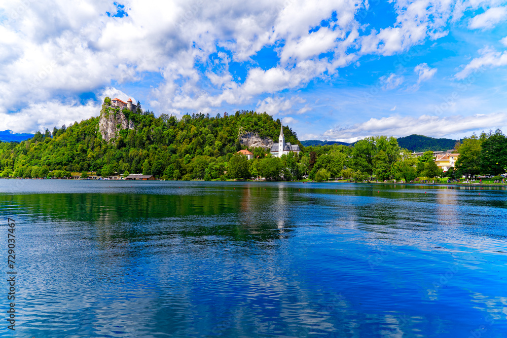 Obraz premium Scenic view of Lake Bled with beautiful reflections and castle on a rock and woodland in the background on a blue cloudy summer day. Photo taken August 8th, 2023, Bled, Slovenia.