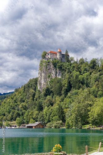 Scenic view of castle on top of rock with Slovenian flag waving at lakeshore of Slovenian Lake Bled on a cloudy summer day. Photo taken August 8th, 2023, Bled, Slovenia.