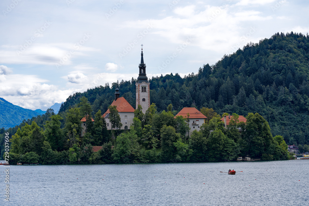Lake Bled with church on an island and woodland in the background on a cloudy summer day. Photo taken August 8th, 2023, Bled, Slovenia.