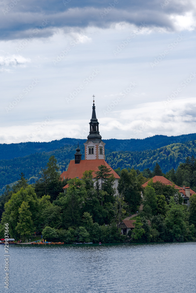 Lake Bled with church on an island and woodland in the background on a cloudy summer day. Photo taken August 8th, 2023, Bled, Slovenia.