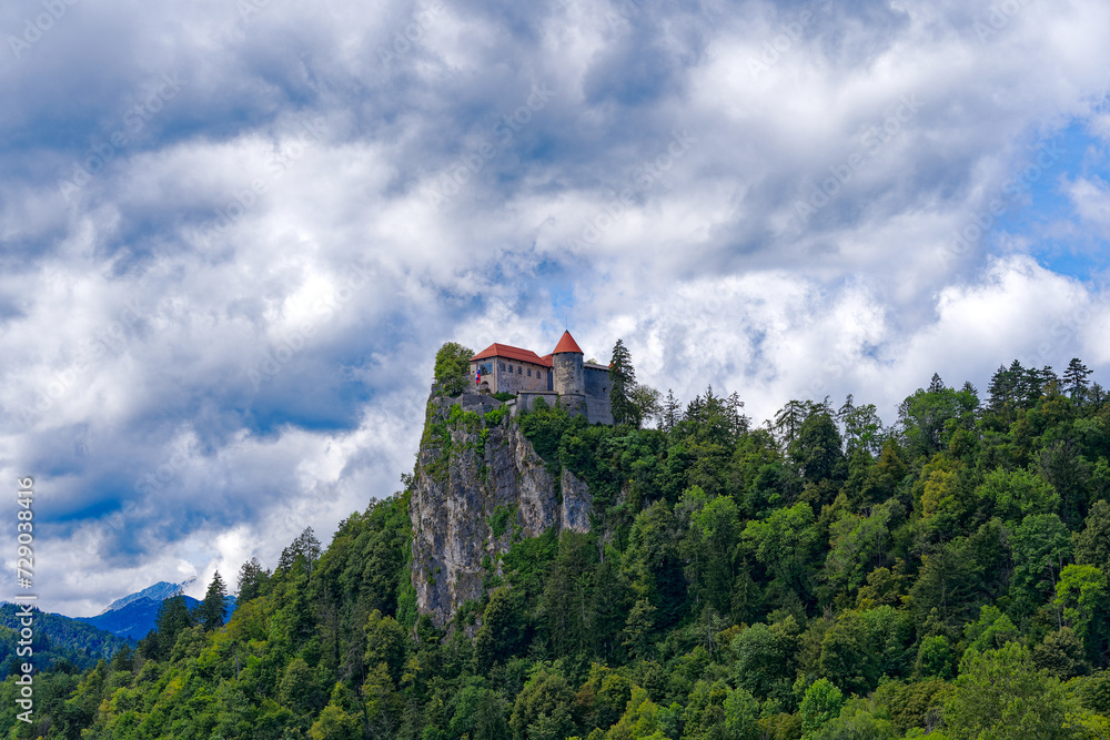 Castle on top of rock with waving Slovenian flag at lakeshore of Slovenian Lake Bled on a blue cloudy summer day. Photo taken August 8th, 2023, Bled, Slovenia.