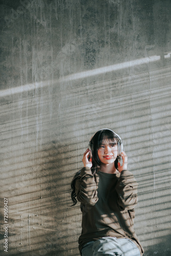 Young Asian woman sitting and listening to music with headphones cement wall backdrop Empty space for creating messages