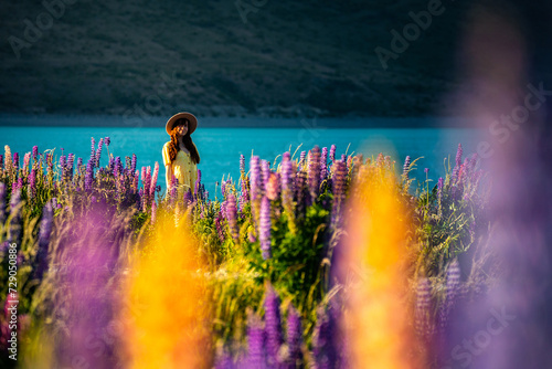 beautiful girl in yellow dress and hat standing on the field of colorful lupins and enjoying the sunset over lake tekapo; unique flowers near mountaineous lake in new zealand, south island, canterbury photo
