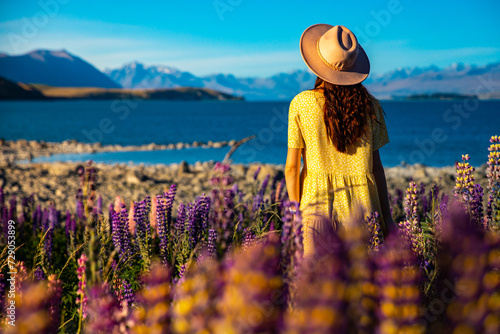 beautiful girl in yellow dress and hat standing on the field of colorful lupins and enjoying the sunset over lake tekapo; unique flowers near mountaineous lake in new zealand, south island, canterbury photo