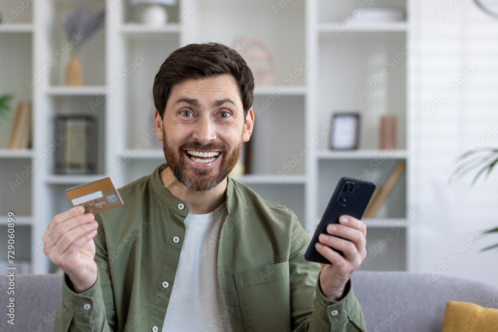 Excited buyer with debit card and mobile phone in pleasant shock from online shopping from home. Amazed male customer getting massive cash back and extra bonuses after making order with shipping. Excited buyer with debit card and mobile phone in pleasant shock from online shopping from home. Amazed male customer getting massive cash back and extra bonuses after making order with shipping.