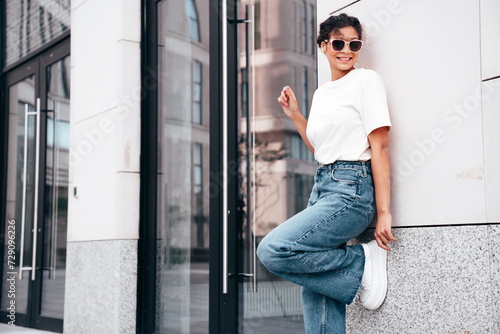 Young beautiful smiling hipster woman in trendy summer white t-shirt and jeans clothes. Carefree woman, posing in the street at sunny day. Positive model outdoors near wall. In sunglasses