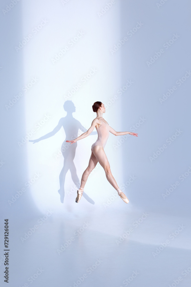 Ballet Beauty. Body-Size Portrait of athlete young woman showcases balletic prowess in pink sport swimsuit and pointe against white backdrop, casting delicate shadow. Concept of grace, athleticism.
