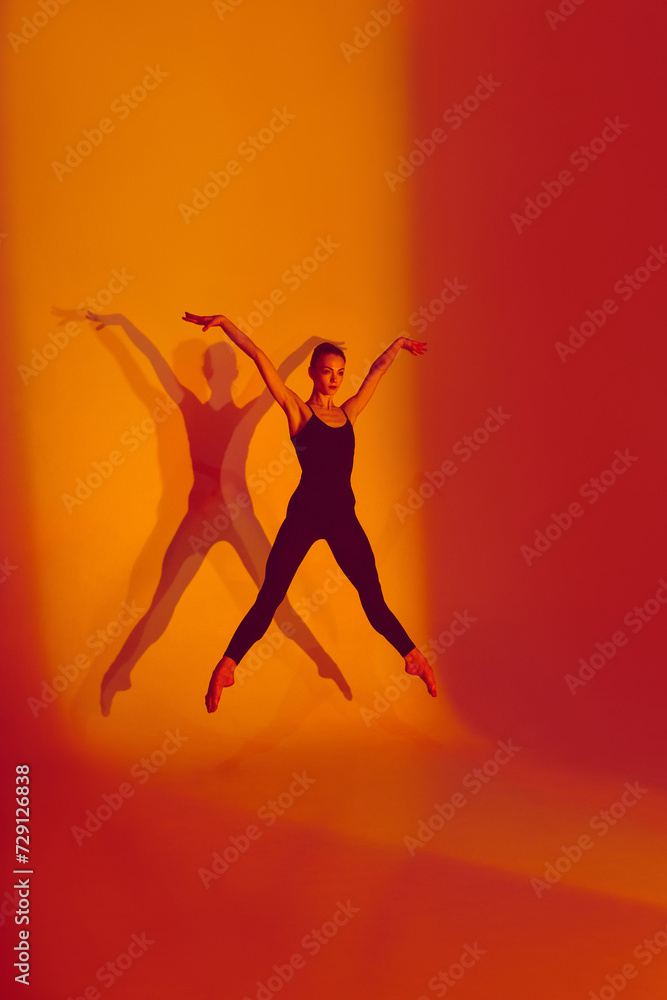 Flexible Elegance. Portrait of graceful dancer poses in black sports overalls, barefoot in neon-lit studio against gradient background with her shadow. Concept of grace, flexibility, athleticism.