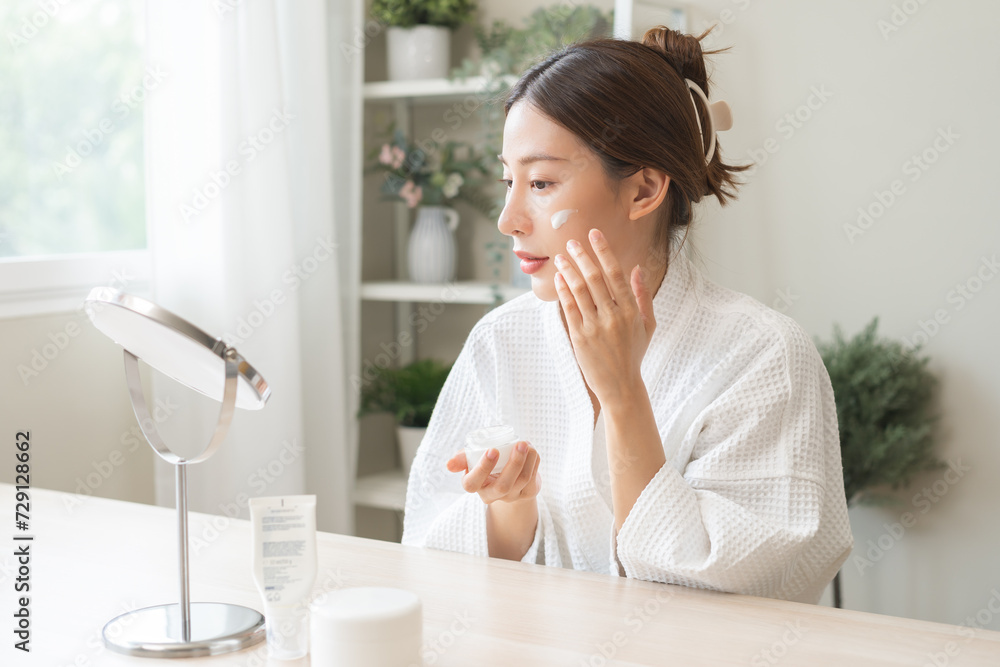 Facial beauty skin care, smile of pretty asian young woman in bathrobe looking at mirror, hand applying moisturizer lotion on her face, holding jar of skin cream before makeup cosmetic routine at home Facial beauty skin care, smile of pretty asian young woman in bathrobe looking at mirror, hand applying moisturizer lotion on her face, holding jar of skin cream before makeup cosmetic routine at home