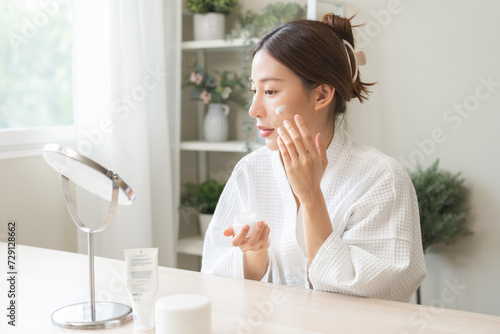 Facial beauty skin care  smile of pretty asian young woman in bathrobe looking at mirror  hand applying moisturizer lotion on her face  holding jar of skin cream before makeup cosmetic routine at home
