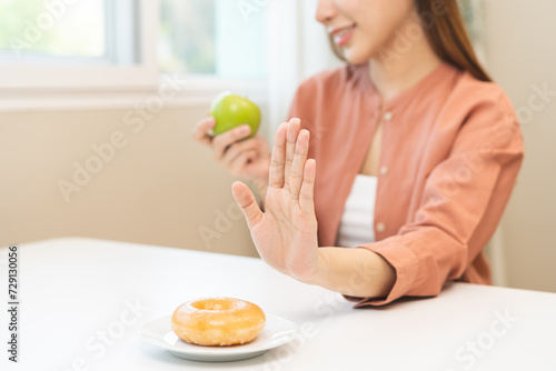 Diet concept, close up young woman, girl using hand push out, stop sweet donut, dessert or junk food on plate at home, choose green apple, eat low fat meal for good health. Female getting weight loss.